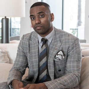 Man wearing plaid suit sitting down on a beige couch wearing a gold metal lapel pin pen