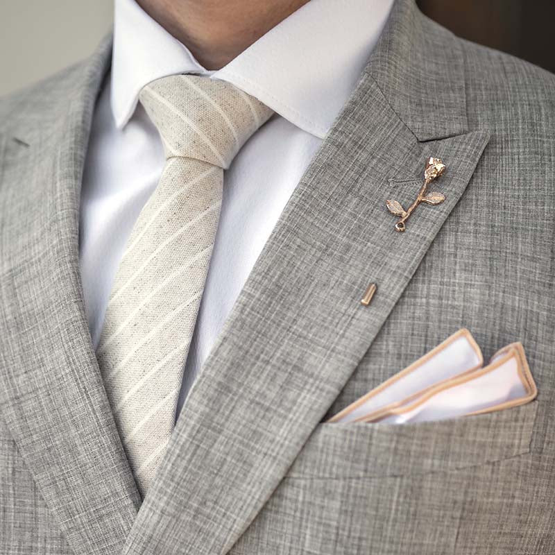 Gray suit with white shirt, light champagne linen striped tie, and decorative lapel pin.