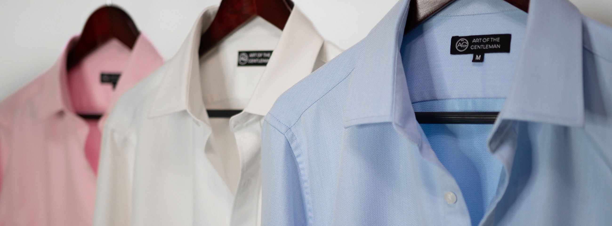 Closeup of dress shirts hanging on a rack. 3 shirts one pink, one white, one blue with Art of The Gentleman Tags