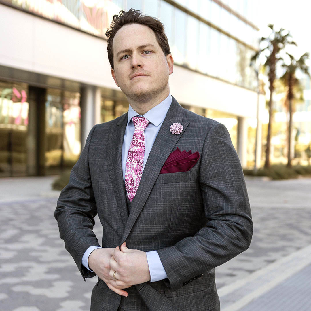 Man wearing a grey plaid business suit with a blue shirt, mauve and burgundy floral tie in an urban environment.