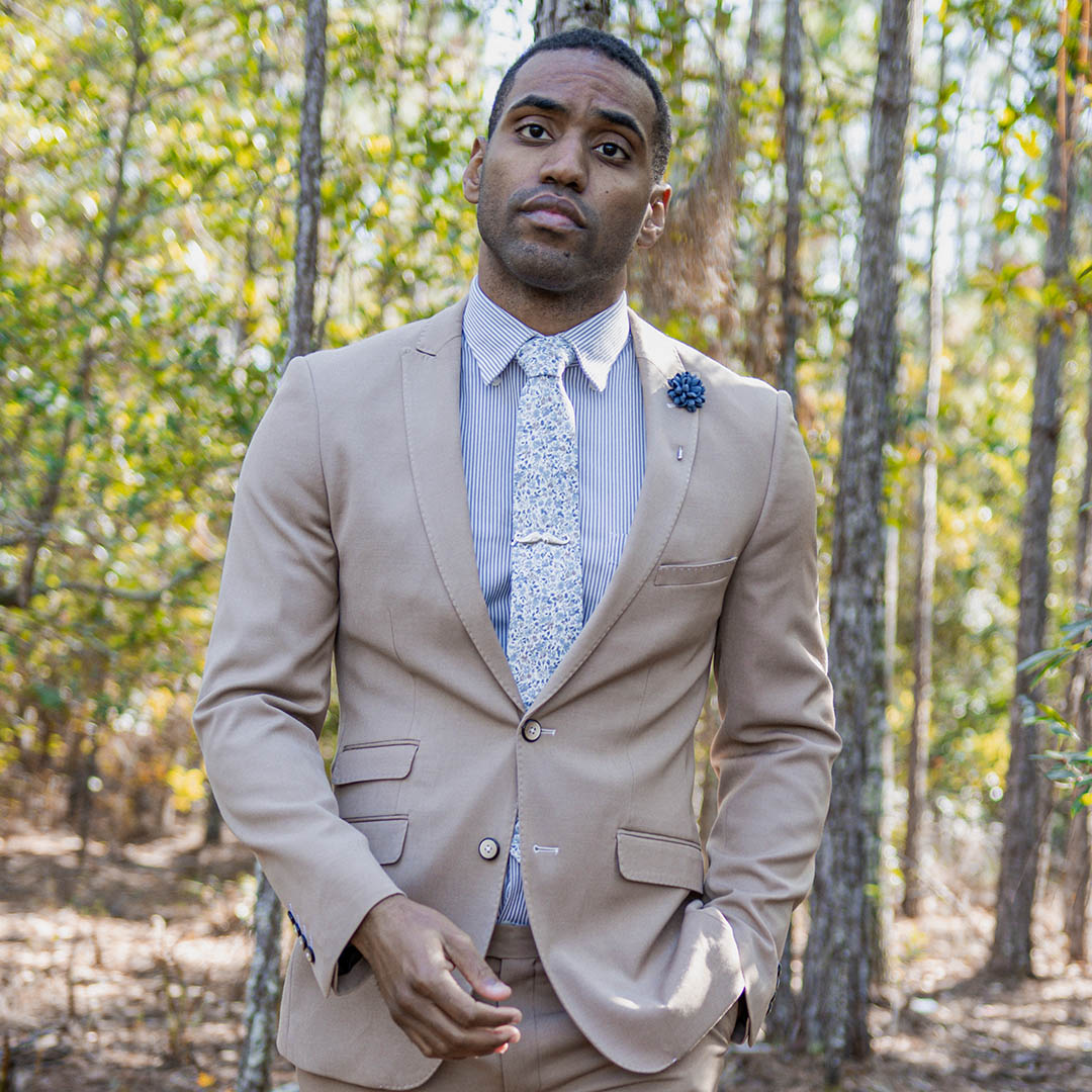 Man wearing a tan suit with a blue striped shirt and floral blue tie in a wooded area