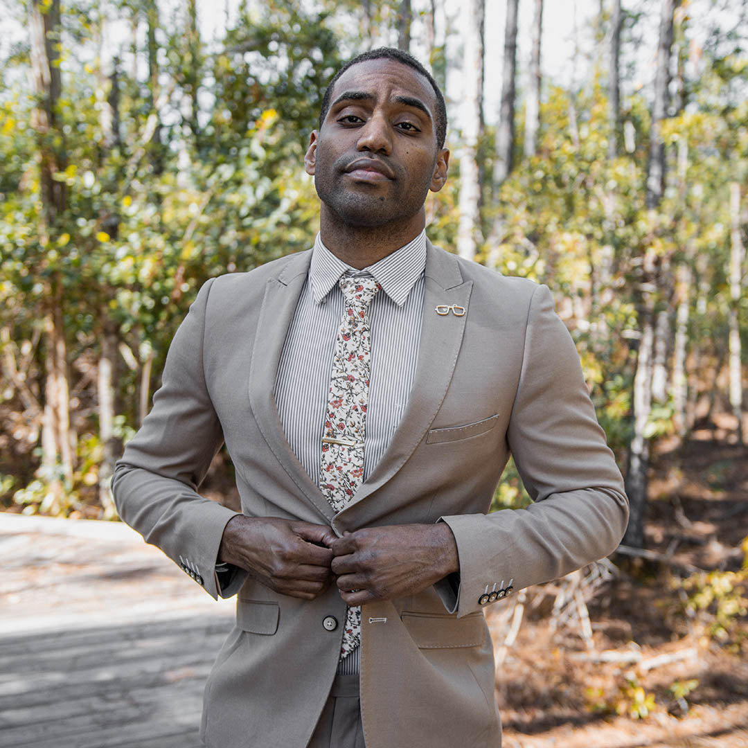 Man wearing a Tan suit with a striped shirt and Floral Cream and Rust Tie in a wooded area