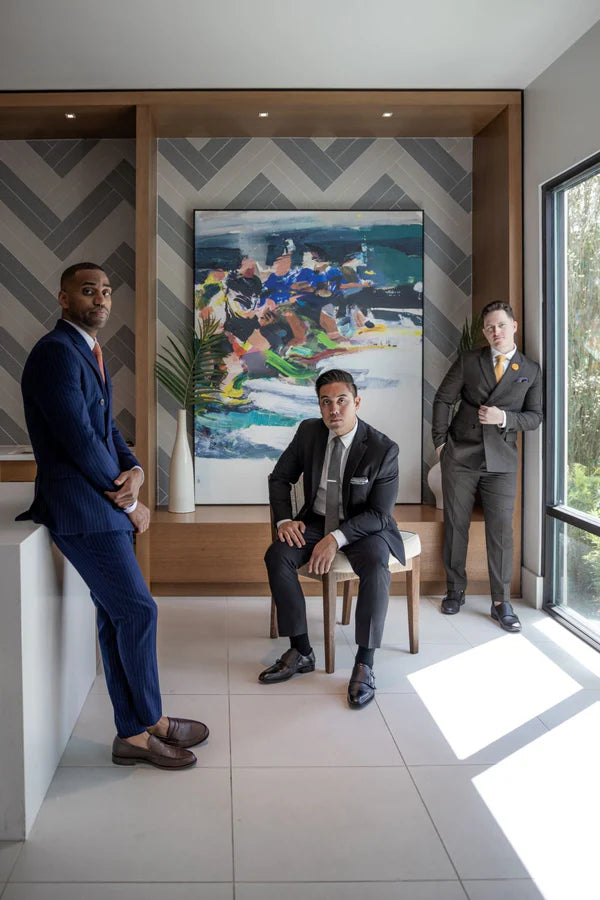 Three men in suits standing in a modern interior setting with a large painting on the wall.