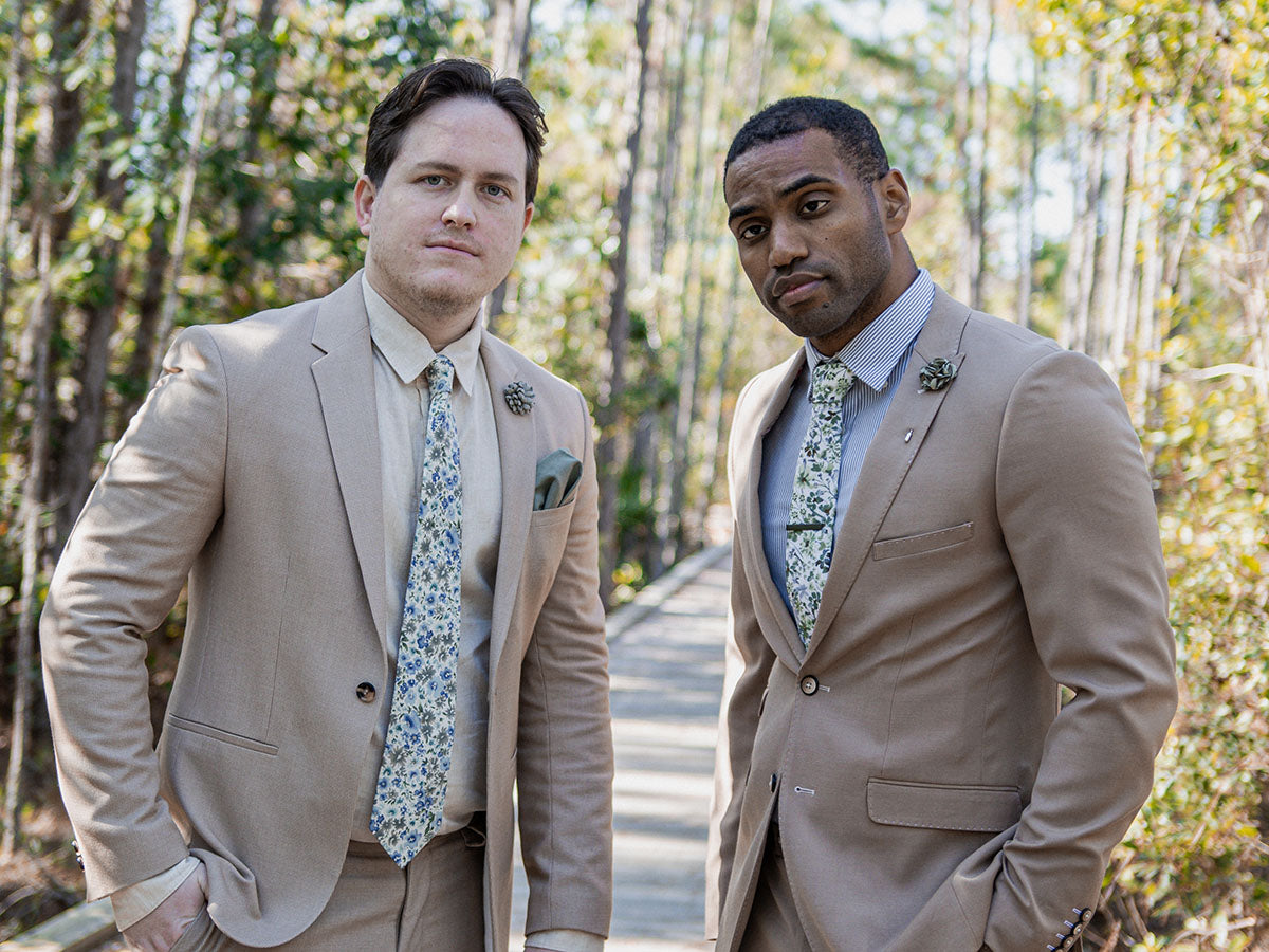 Two men in beige suits with floral ties standing outdoors in a forest.