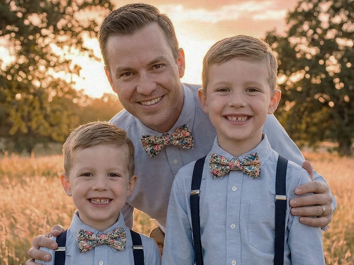 Man and two young boys wearing floral bow ties and suspenders standing in a field with a warm, blurred background.