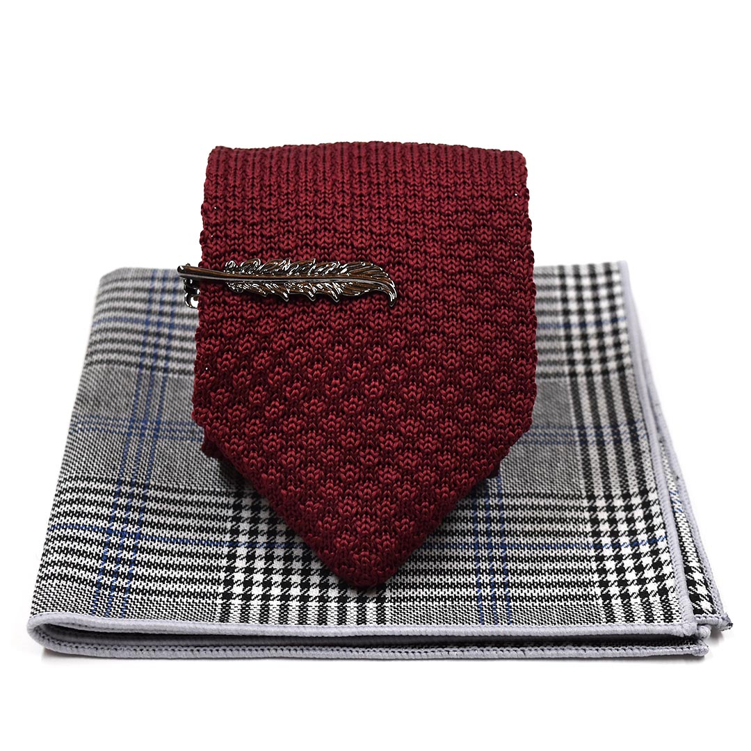 A burgundy knit necktie with a silver feather tie clip, displayed on top of a gray and black plaid pocket square against a white background.