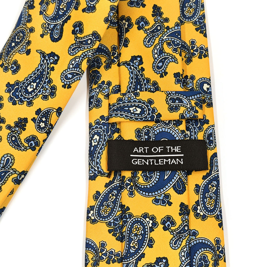 Yellow tie with blue paisley pattern and 'Art of the Gentleman' label on a white background