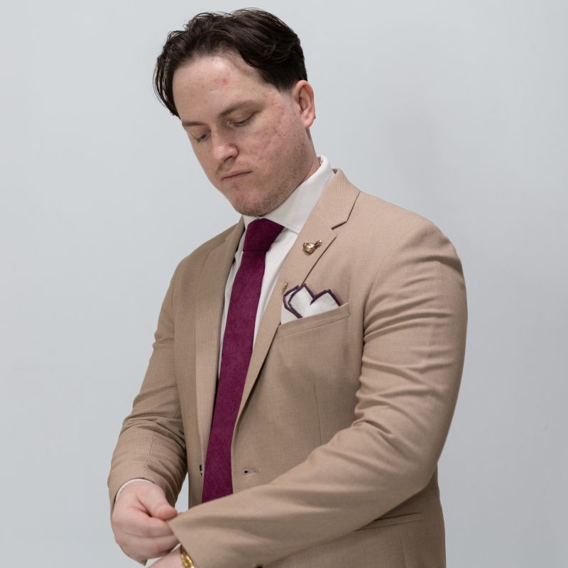 Man in a beige suit with a red tie and white pocket square against a plain background