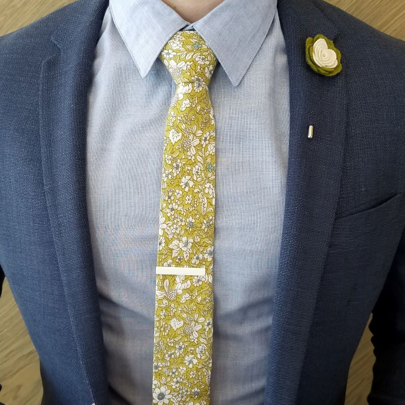 Floral Snow White Olive lapel pin worn with a navy suit jacket, a light blue shirt, a necktie and a tie bar